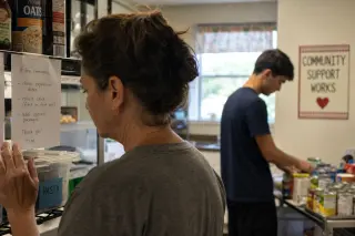 A woman reads a note in a community kitchen as a man organizes shelves, both engaged in quiet volunteer tasks under gentle daylight.