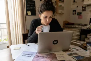 Young woman in a cozy kitchen, focused on her laptop and charity-related notes, with gentle sunlight creating a warm, authentic atmosphere.