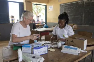 Retired woman and young local woman organizing medical supplies together at a sunlit classroom desk, captured candidly with a gentle, natural ambiance.