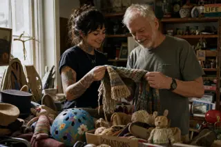 Two people in a thrift store sort through quirky donated items, sharing a subtle smile, with soft daylight creating a warm, candid atmosphere.