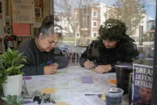 Two people collaborating on a handwritten map at a community center table, sunlight streaming through the window and urban buildings in the background.