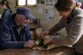 A woman wraps vegetables for an elderly man in a rural community hall, with supplies in the background and soft daylight illuminating the scene.