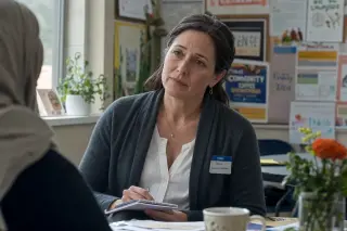 A woman at a table in a sunlit community room, holding a notepad and listening attentively, with soft daylight highlighting a moment of reflection.