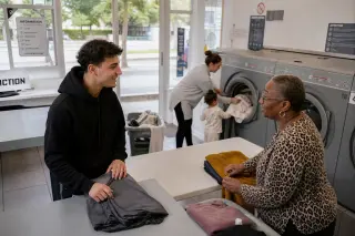 Three people from diverse backgrounds helping each other with laundry in a sunlit urban laundromat, captured candidly in a warm and genuine moment of local solidarity.